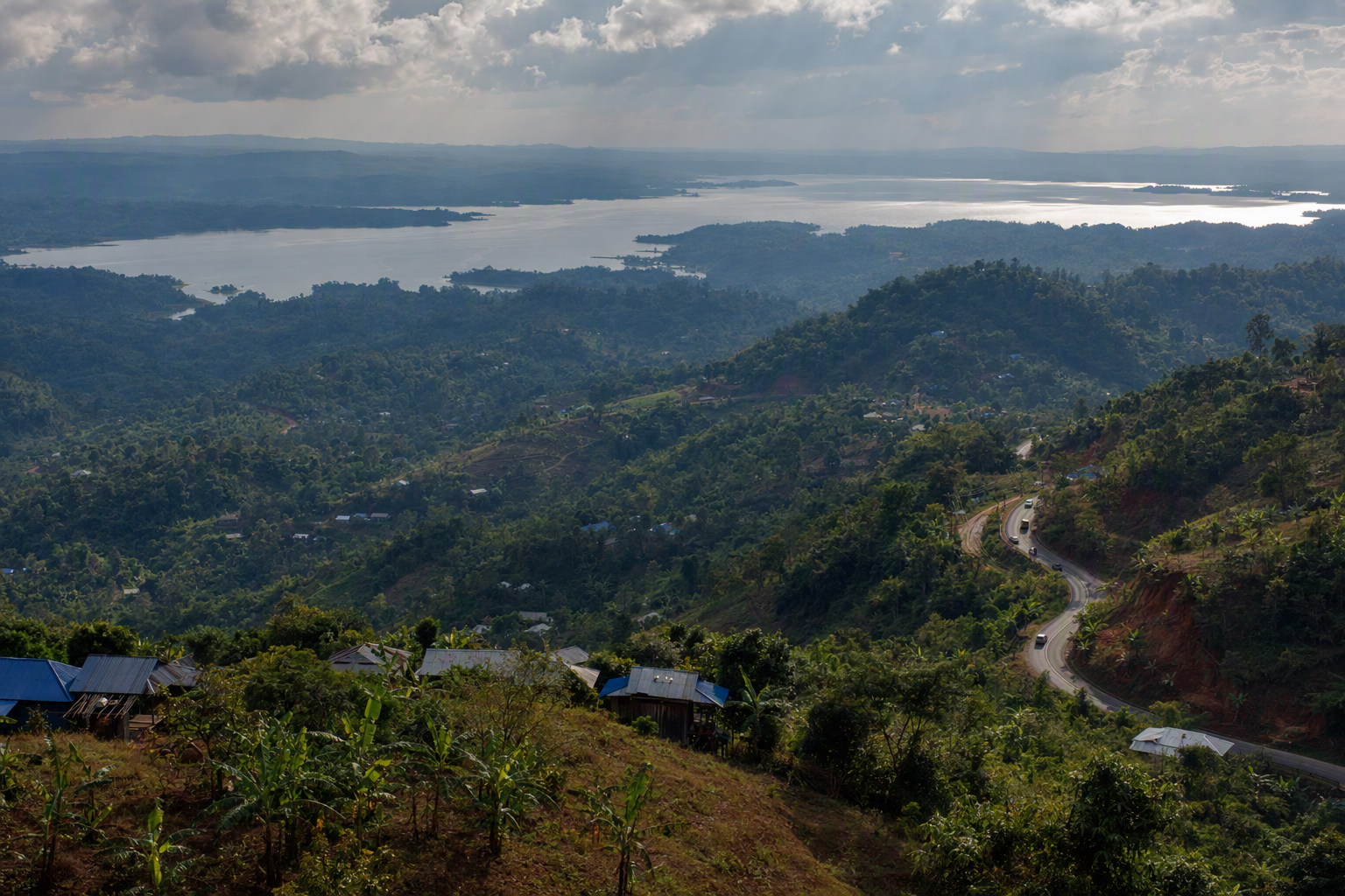 Wide view across forested ridges, water, roads, and small settlements in the Chittagong Hill Tracts