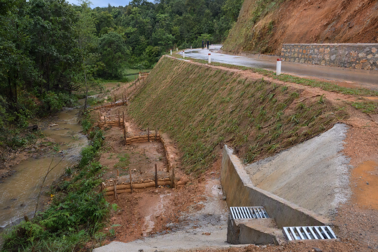 A hill road under routine maintenance with a drained cut slope, retaining work, sediment traps, vegetation, and a stream below