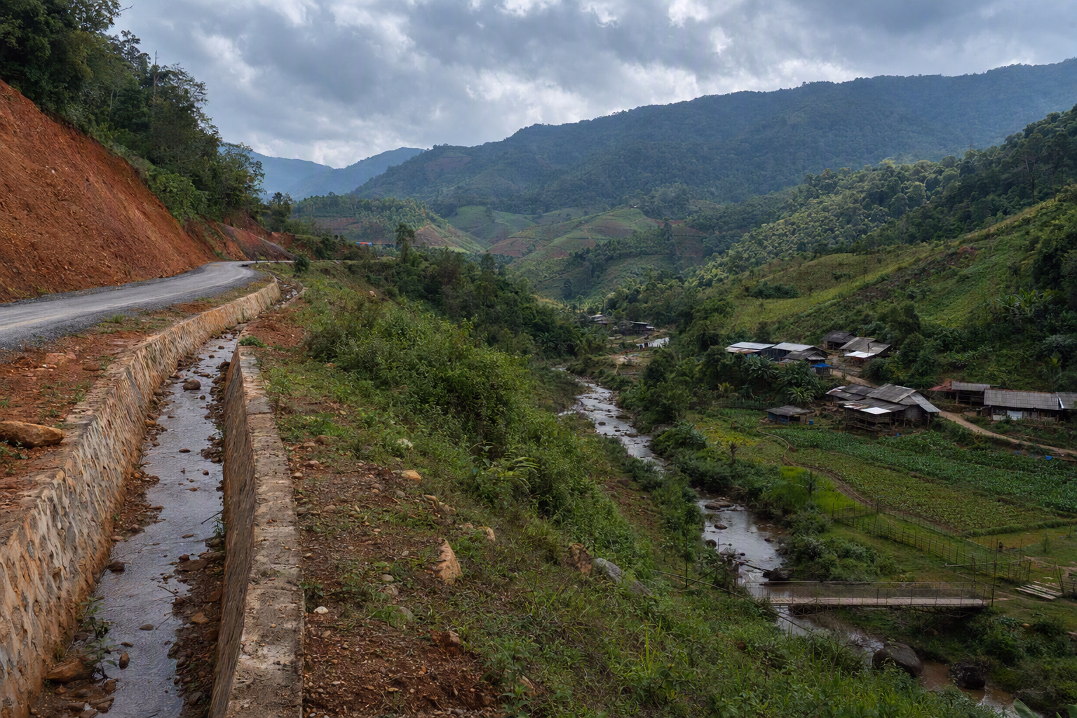A broad hill watershed with a road cut, drainage channel, mixed forest, cultivated slopes, a stream, and downstream settlement visible in one frame