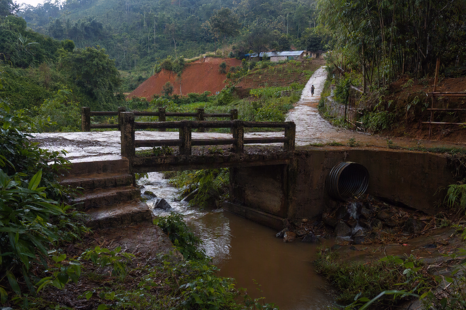 A rain-fed hill stream passing beneath a weathered footbridge, with layered ridges, exposed soil, drainage channels, and mixed vegetation visible together