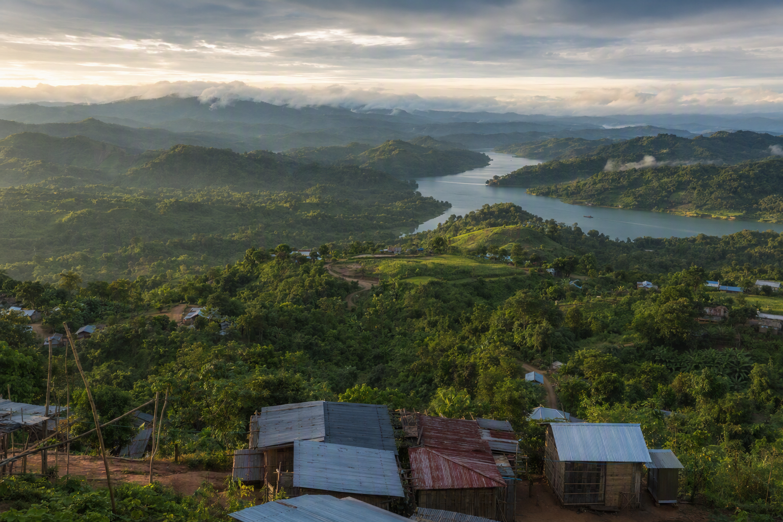 Layered forested ridges descending toward a broad waterway, with small distant settlements and boats under monsoon mist