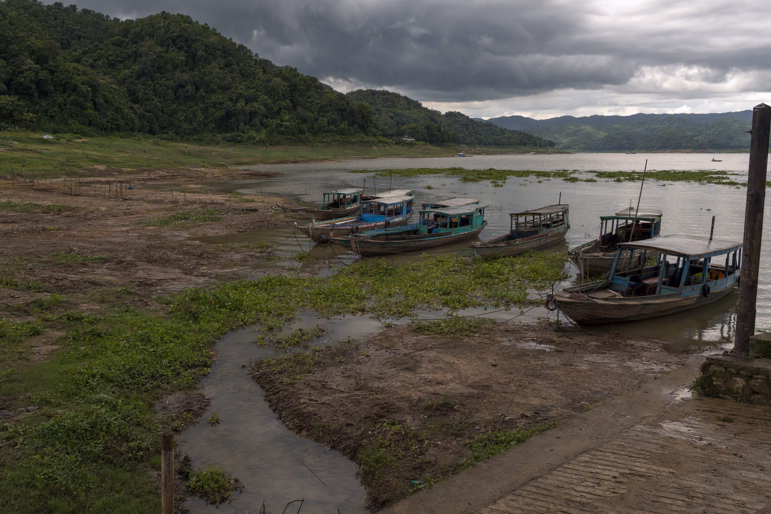 A generalized reservoir inlet with an exposed seasonal shoreline, moored passenger boats, floating vegetation, rain clouds, and forested slopes